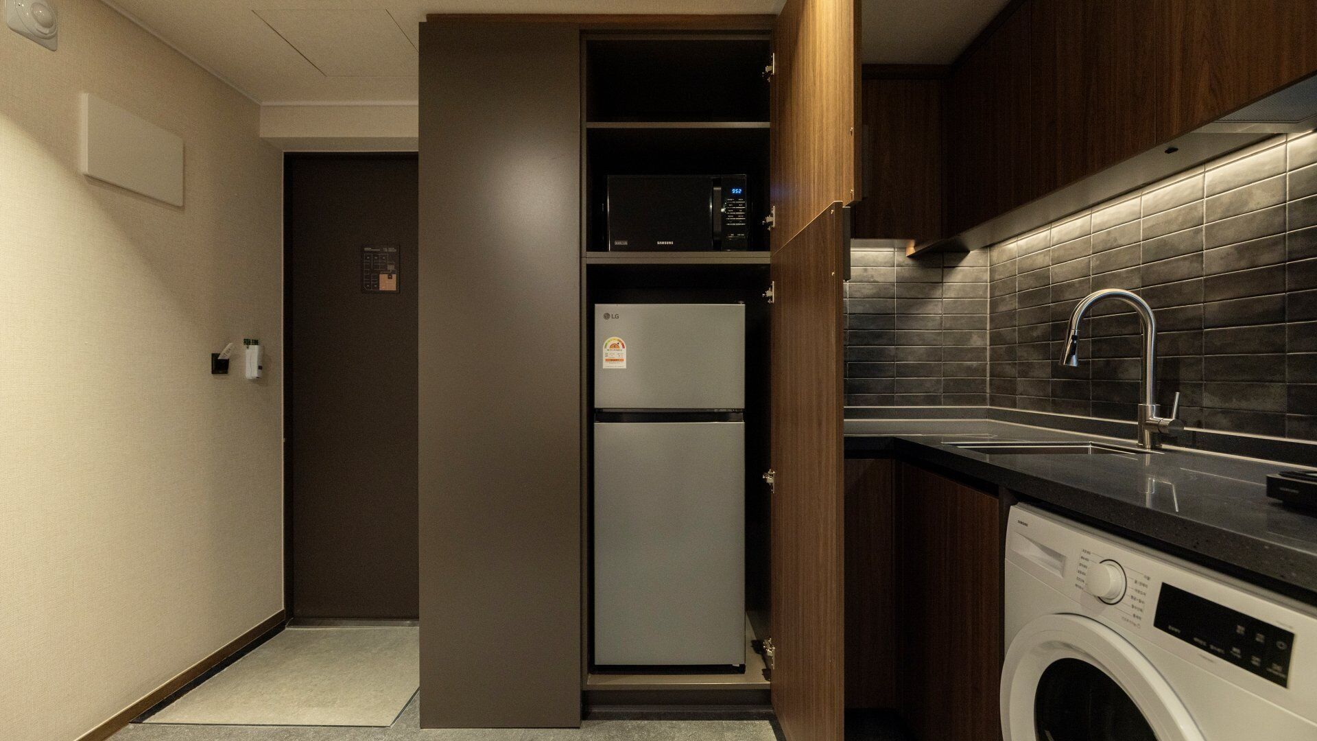 Una cocina compacta con lavadora integrada, un lavamanos de acero inoxidable y un salpicadero de azulejos oscuros.