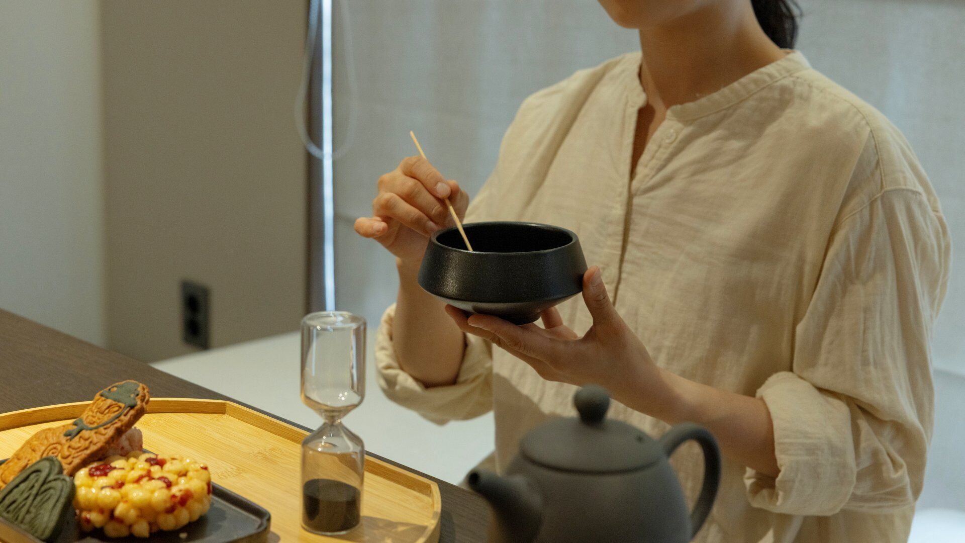 A person holding a black bowl with chopsticks, a tray with food items, and a teapot on a table.