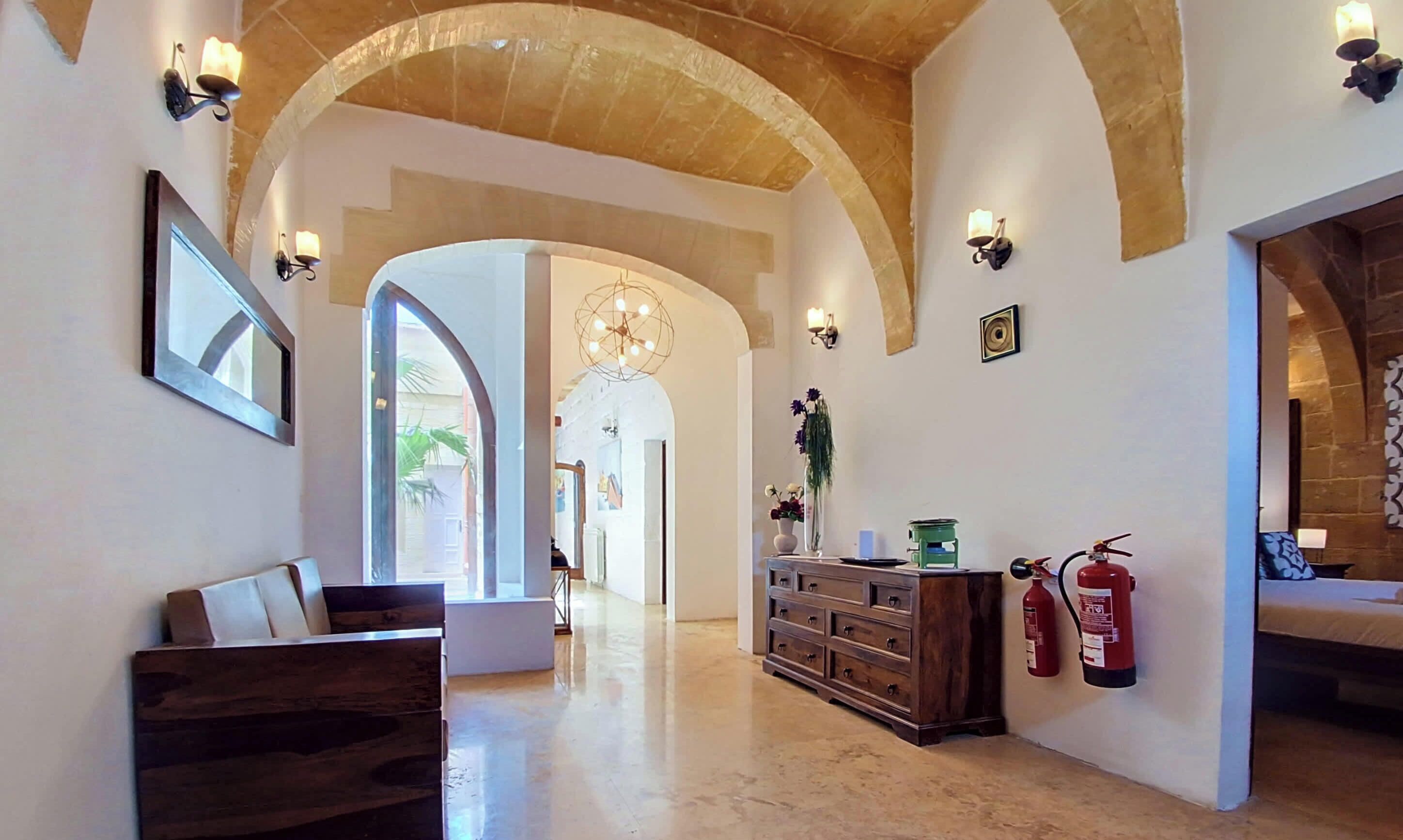 A hallway with arched ceilings, a wooden bench, a dresser, wall sconces, and a fire extinguisher.