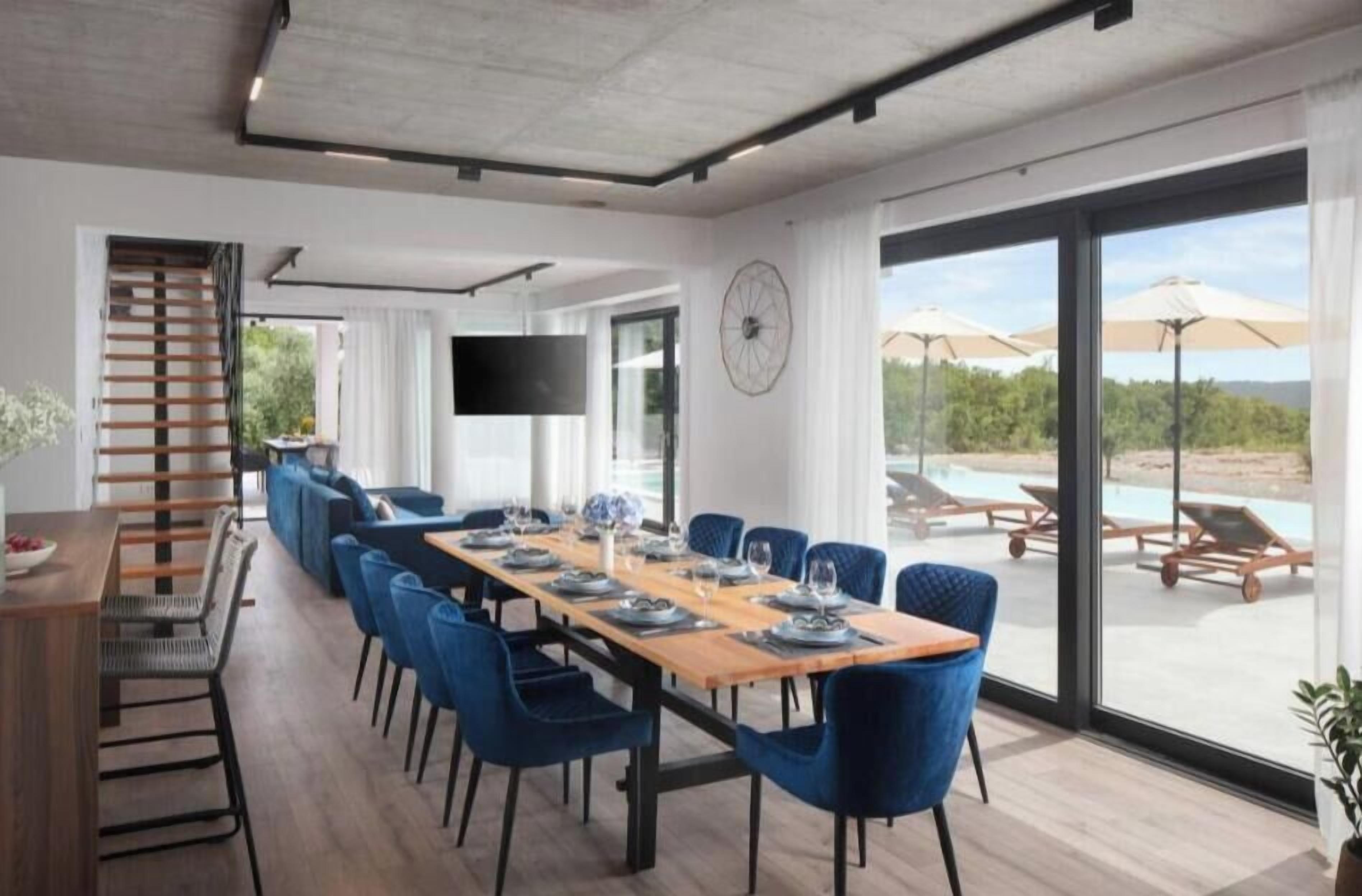 Een moderne eetruimte met een lange tafel voor zes personen, blauwe stoelen, een houten bank en een televisie aan de muur.