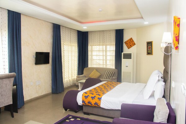 A hotel room with a bed, a sofa, a TV, a window with curtains, and a wall-mounted air conditioner.