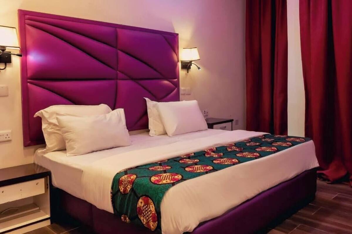 A hotel room with a large bed, a headboard, two bedside lamps, a nightstand, and a window with red curtains.