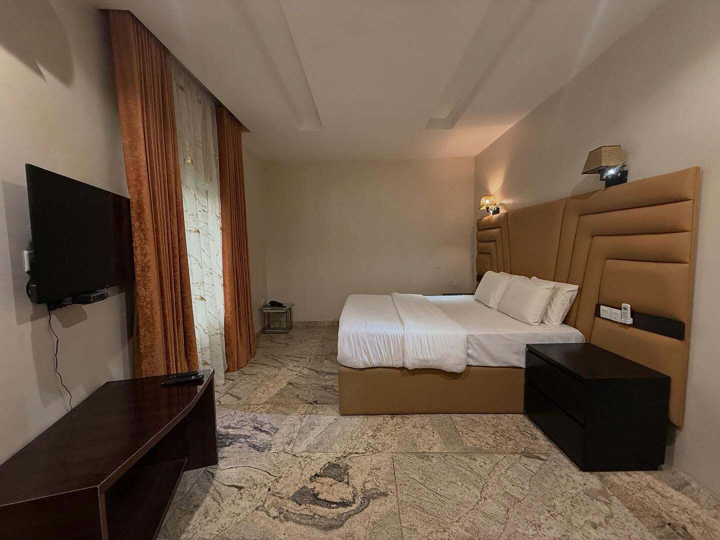 Una habitación de hotel con una cama, televisión, mesita de noche, lámpara y una pequeña mesa.