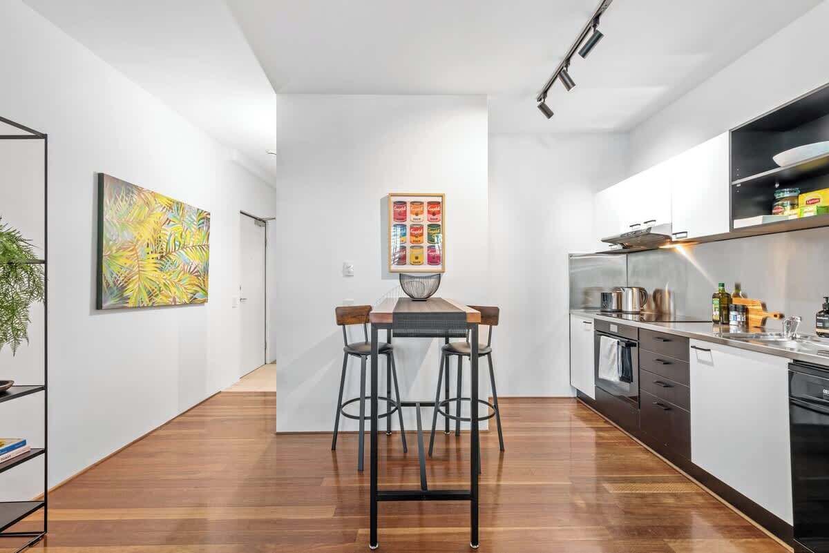 A modern kitchen with a wooden table and bar stools, a painting on the wall, and a kitchen island with appliances.
