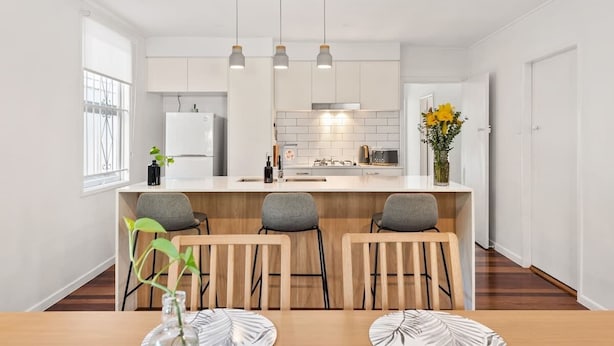 Una cocina moderna con armarios blancos, una isla central con taburetes y luminarias colgantes, con vistas al área de comedor contigua.