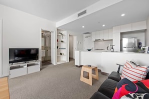 A modern living room with a TV, a small stool, a sofa, and a kitchen area with white cabinets and appliances. - Captivating Convenience! 1bed/1bath/1car/views (Brisbane)