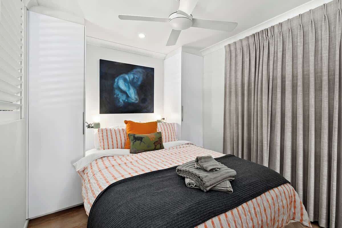 A modern bedroom with a bed, a ceiling fan, a large window with blinds, and a painting on the wall.