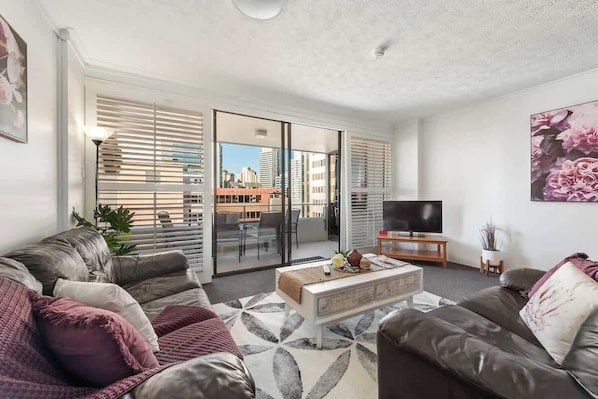 A modern living room with a sofa, coffee table, and TV, with a view of the city through large windows. - Amazing Astor ~ 2bed, 2bath, 1car, Pool! (Brisbane)