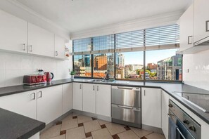 A modern kitchen with white cabinets, stainless steel appliances, a large window with blinds, and a city view. - Amazing Astor ~ 2bed, 2bath, 1car, Pool! (Brisbane)