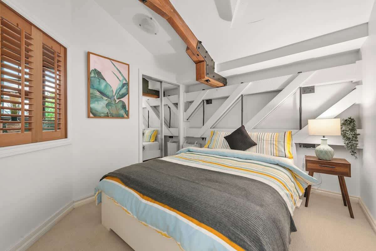 A bedroom with a bed, a window with blinds, a painting, a lamp, and a plant.