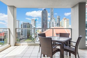 Un balcón con una mesa de vidrio y sillas de mimbre, con vistas a un paisaje urbano con rascacielos.