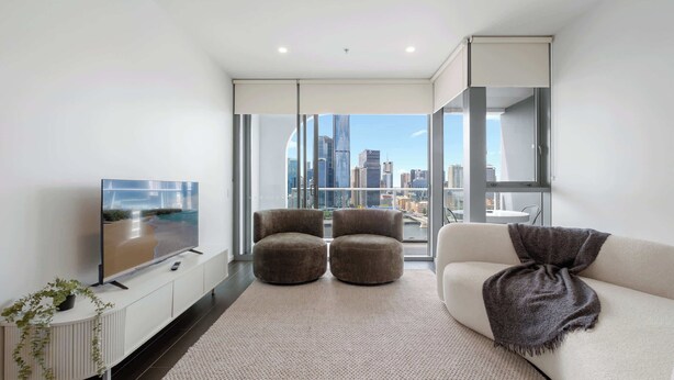A modern living room with a large TV, two ottomans, a sofa, and a city view through floor-to-ceiling windows.