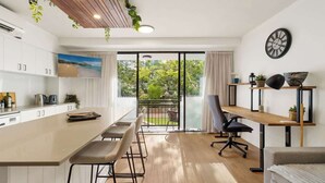 Eine moderne Küche mit einer großen Kochinsel, einem Essbereich mit Stühlen, einem Holztisch und Blick durch Glastüren auf einen Außenbalkon.