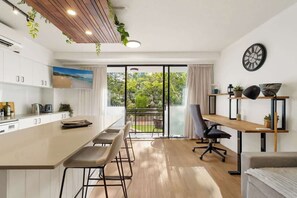 Eine moderne Küche mit einer großen Kochinsel, einem Essbereich mit Stühlen, einem Holztisch und Blick durch Glastüren auf einen Außenbalkon.