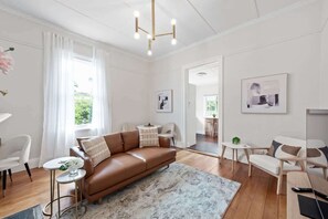 A modern living room with a brown leather sofa, a round coffee table, and a large window with white curtains. - Art Deco With Modern Twist! 1bed-1bath-1car-new Farm (Brisbane)