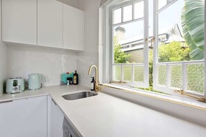 A kitchen with white cabinets, a sink, a window with a view of greenery, and a teal-colored appliance. - Art Deco With Modern Twist! 1bed-1bath-1car-new Farm (Brisbane)