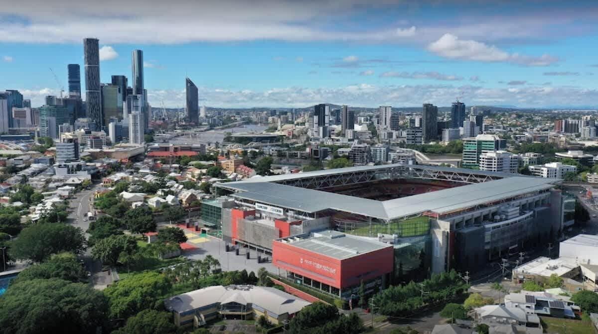 A stadium with a large central field surrounded by modern buildings, including a distinctive red building with a glass facade.