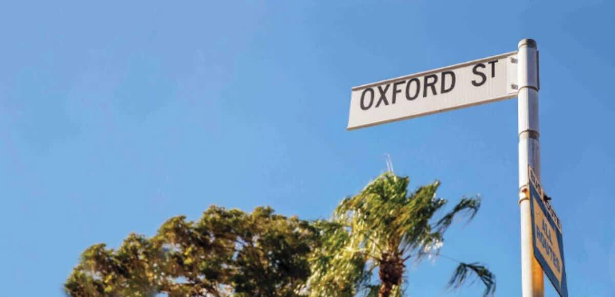 A street sign for 'OXFORD ST' with a clear blue sky and trees in the background.