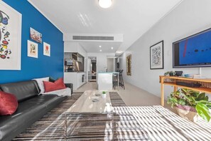 Ein modernes Wohnzimmer mit einer schwarzen Ledersofa, einem Couchtisch und einem Fernseher rechts. Im Hintergrund befindet sich eine Küche mit einem Essbereich.