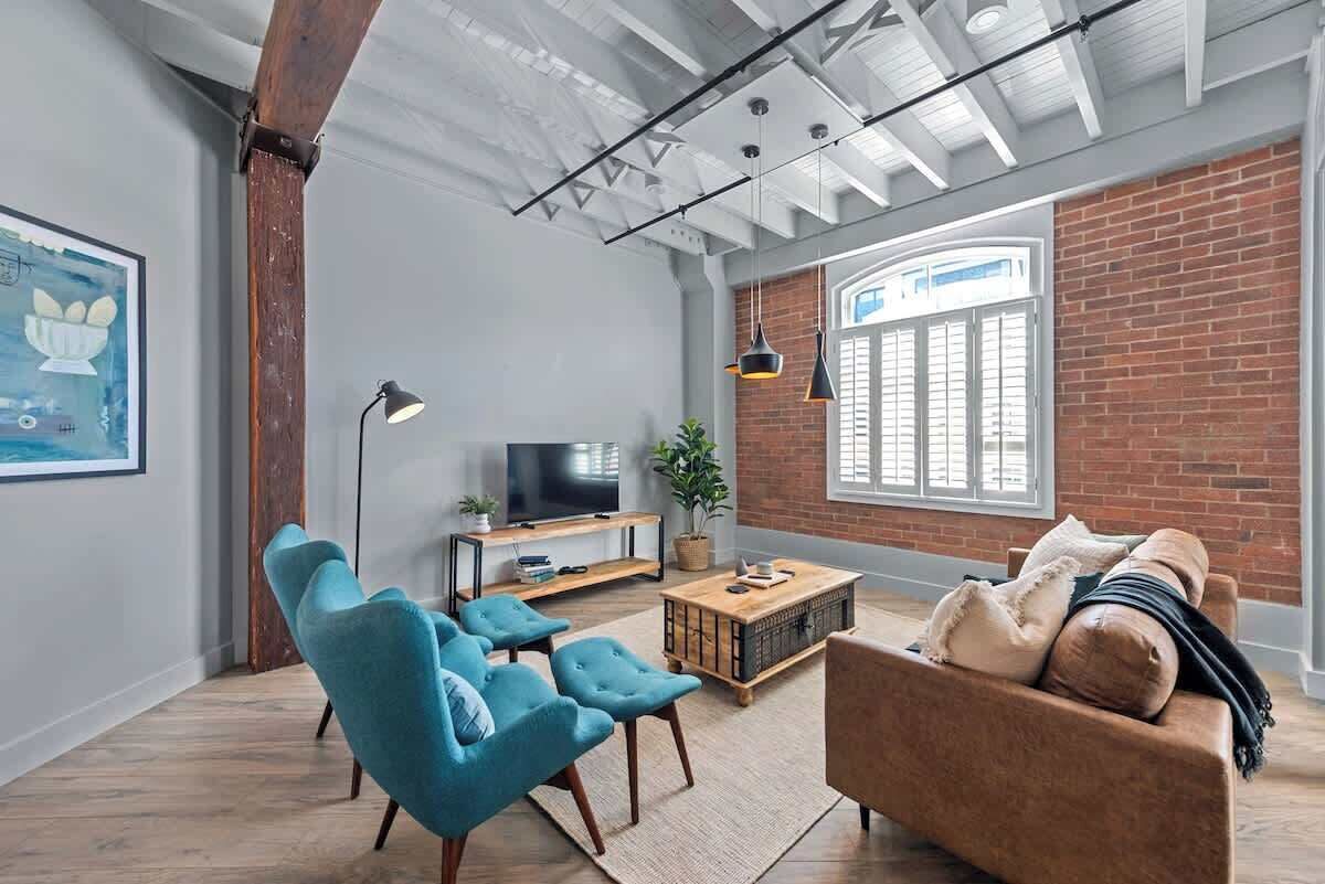 Una sala de estar moderna con televisor, sillones azules, un sofá marrón, una mesa de centro de madera y una pared de ladrillo.