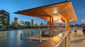 A waterfront walkway with a covered seating area, a view of the city skyline, and a bridge in the background. - Luxurious Woolstore Living! ~teneriffe (Brisbane)