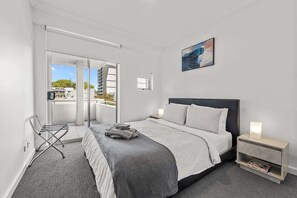 Quarto de hotel moderno com cama, mesa de cabeceira, luminária na parede, janela com vista e porta para área externa.