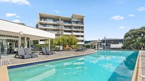 Una piscina con sillas de descanso, un edificio moderno con balcones y un cielo despejado.