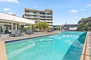 Piscina com espreguiçadeiras, um edifício moderno com varandas e céu claro.