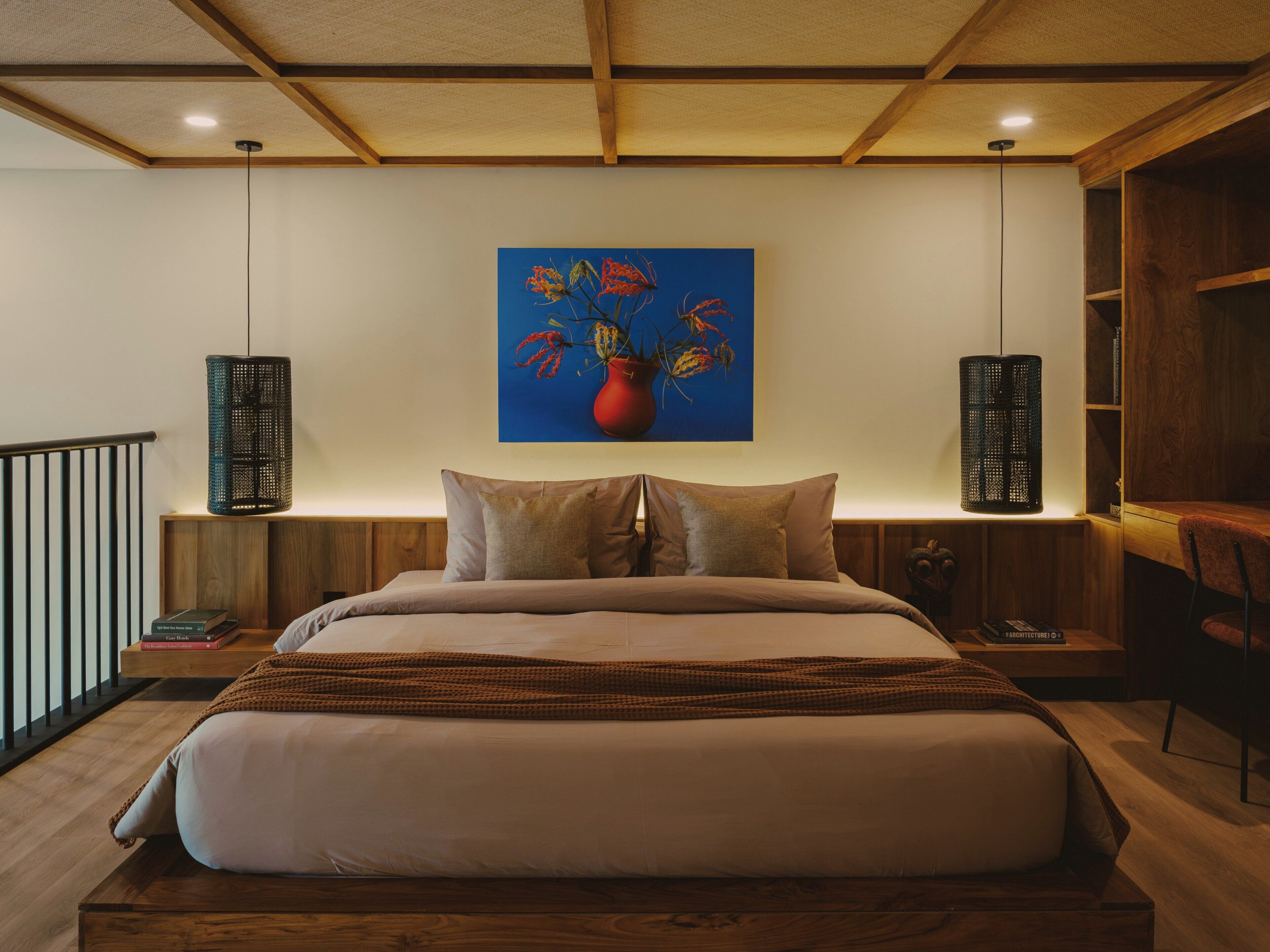 Quarto com cama grande, cabeceira de madeira, pintura de flores, duas luminárias fixadas na parede e mesa de cabeceira de madeira com livros.