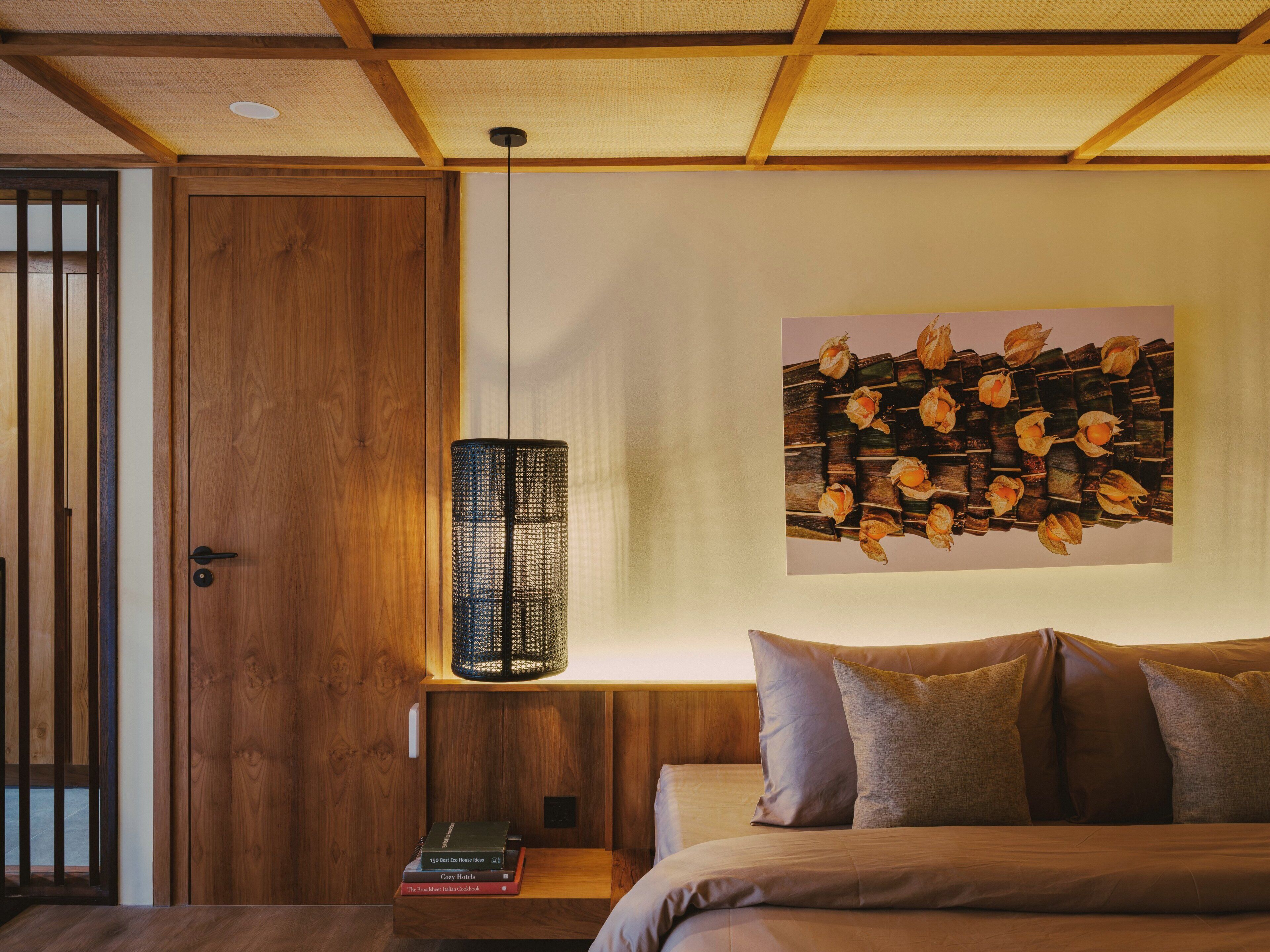 A room with a wooden door, a hanging light fixture, a wall-mounted artwork, a bed with pillows, and a small table with books.