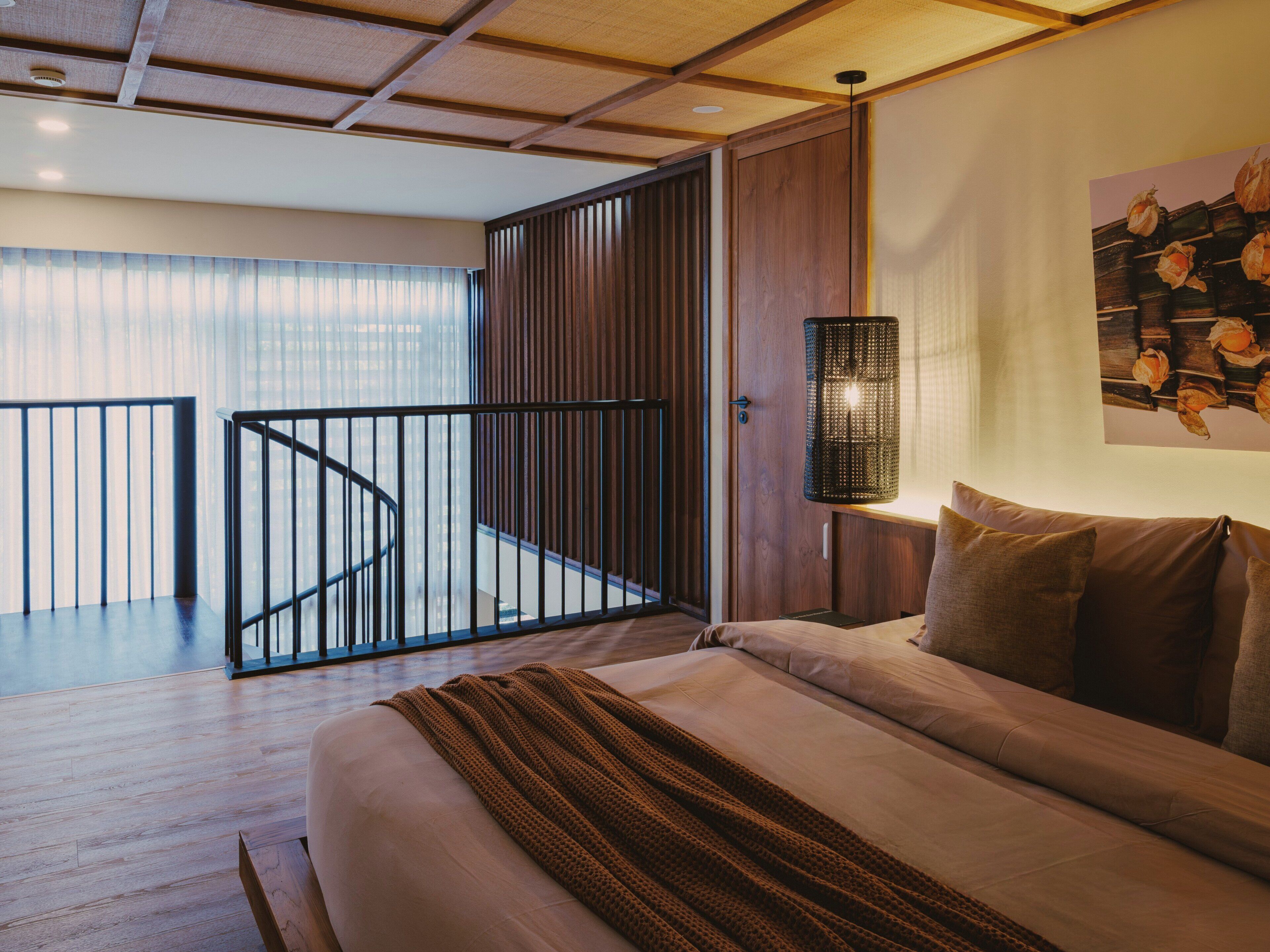 Quarto moderno com cama, piso de madeira, corrimão preto, luminária fixada na parede e um quadro.