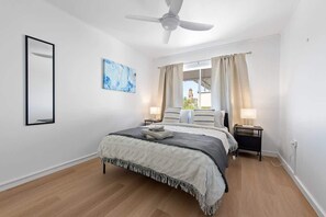 Ein Schlafzimmer mit einem Bett, Deckenventilator, einem Fenster mit Vorhängen, einem Nachttisch und einem an der Wand befestigten Spiegel.