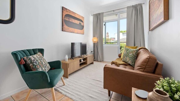 A living room with a sofa, a green armchair, a TV on a wooden stand, and a window with curtains. - New Farm Nest! 2bed/1bath/2car/balcony (Brisbane)