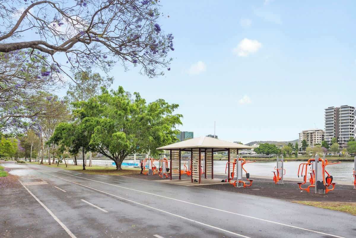 A park with exercise equipment, a covered seating area, and a road with trees and buildings in the background.