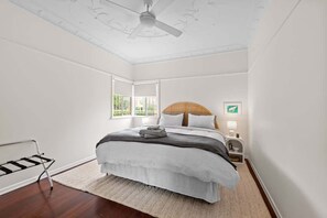 Ein Schlafzimmer mit Bett, Deckenventilator, Nachttisch mit Lampe, Fenster und Holz-Kopfteil.
