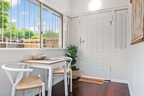 Ein Zimmer mit Schreibtisch, zwei Stühlen, einem Blumentopf, einer Tür und einem Fenster mit Blick auf Grünfläche.