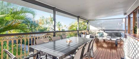 A covered outdoor dining area with a table and chairs, a grill, and a view of the surrounding greenery.