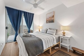 Ein Schlafzimmer mit Bett, Nachttischen, Deckenventilator und Blick durch ein mit Vorhängen versehenes Fenster.