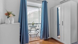 Une pièce avec un plancher en bois, une armoire blanche, une petite table ronde avec des chaises et une fenêtre ornée de rideaux bleus.