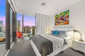 Ein modernes Hotelzimmer mit einem großen Bett, einem roten Sessel, Blick auf die Stadt und einem farbenfrohen Gemälde an der Wand.