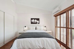 Ein Schlafzimmer mit Bett, Nachttischen, einer Wand-Klimaanlage und einem Fenster mit Jalousien.