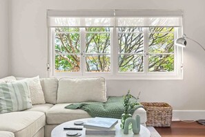 Ein Wohnzimmer mit einem beigefarbenen Sofa, einem runden Couchtisch mit Büchern und einer Fernbedienung, einer grünen Strickdecke, einer Vase mit einer Pflanze und einem Fenster mit Blick auf Bäume.