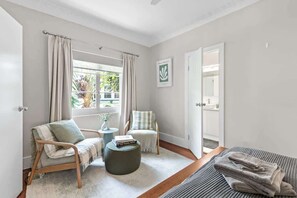 Ein Zimmer mit einem Bett, einem Sessel, einem kleinen Tisch, einem Fenster mit Vorhängen und einer Tür zu einem anderen Raum.
