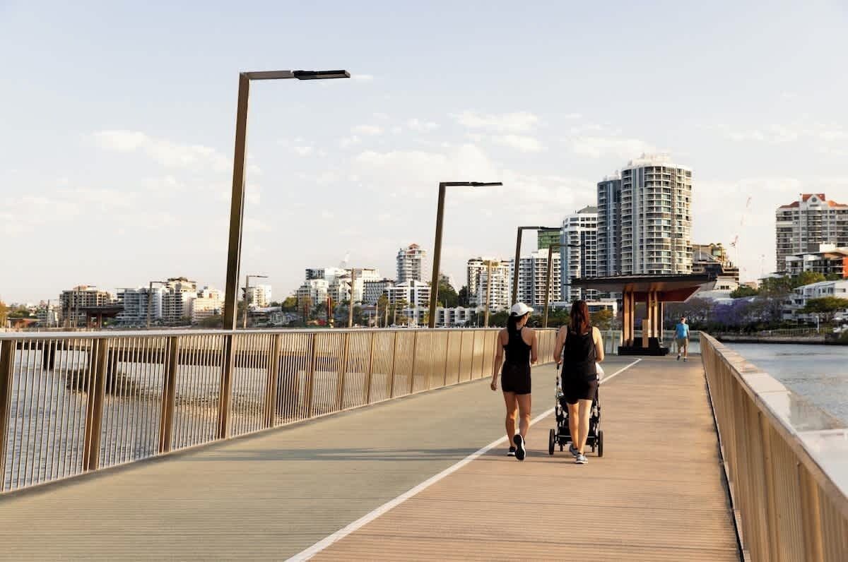 Uma ponte para pedestres com guarda-corpo, céu claro e edifícios ao fundo.