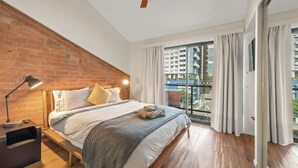 Un dormitorio con pared de ladrillo, cama con colcha a rayas, mesita de noche, ventilador de techo y vista a un paisaje urbano a través de amplias ventanas.