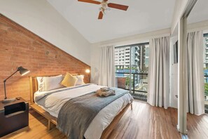 Quarto com parede de tijolos, cama com colcha listrada, mesa de cabeceira, ventilador de teto e vista para a paisagem urbana através de janelas amplas.