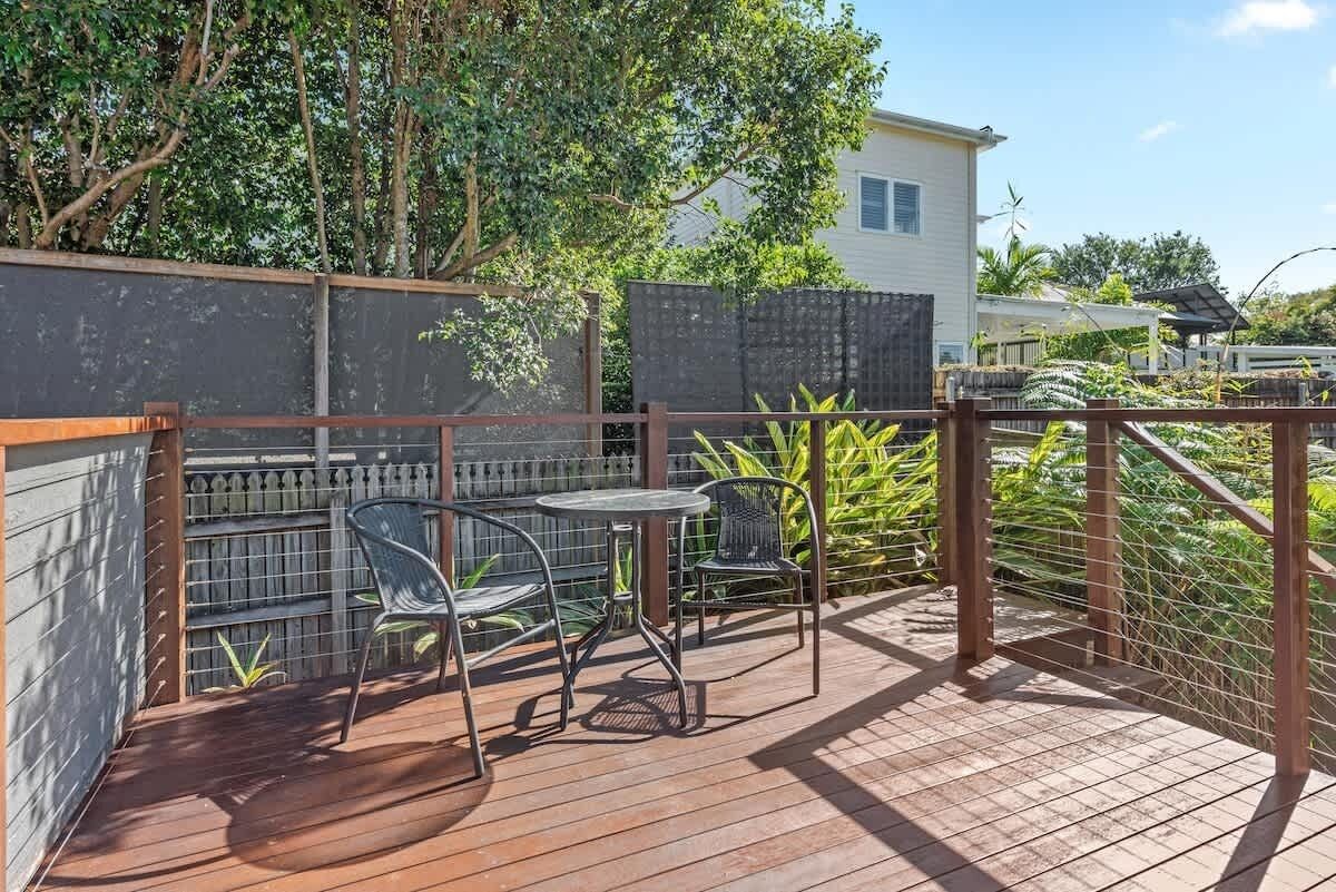 Una terraza de madera con sillas y una mesa de metal, rodeada por una cerca y follaje.