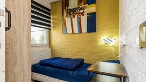 Um quarto pequeno com uma cama, uma mesa de madeira, uma luminária na parede e uma janela.