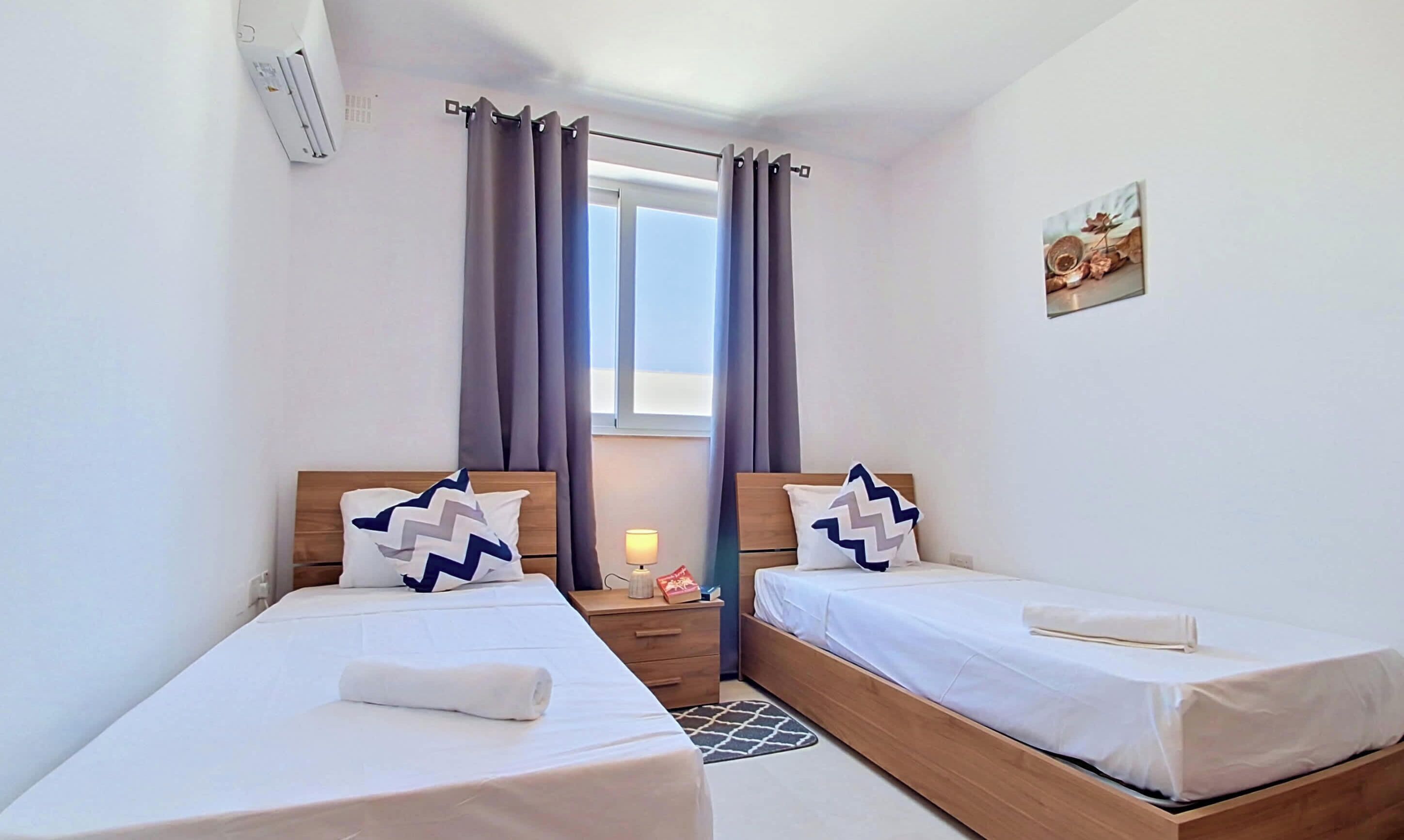 A bedroom with two beds, a window with curtains, a bedside lamp, and a wall-mounted air conditioner.