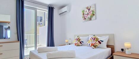 A hotel room with a bed, a nightstand, a lamp, a wall-mounted air conditioner, a window with curtains, and a balcony.
